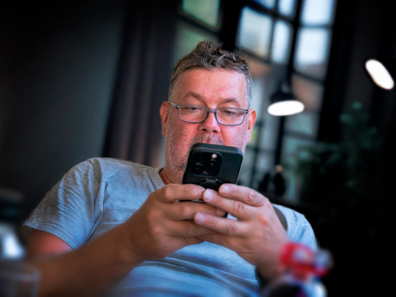 Mario Hambsch sitzt entspannt im Studio, blickt konzentriert aufs Smartphone, unscharfe Flaschen im Vordergrund, Studiolichter im Hintergrund