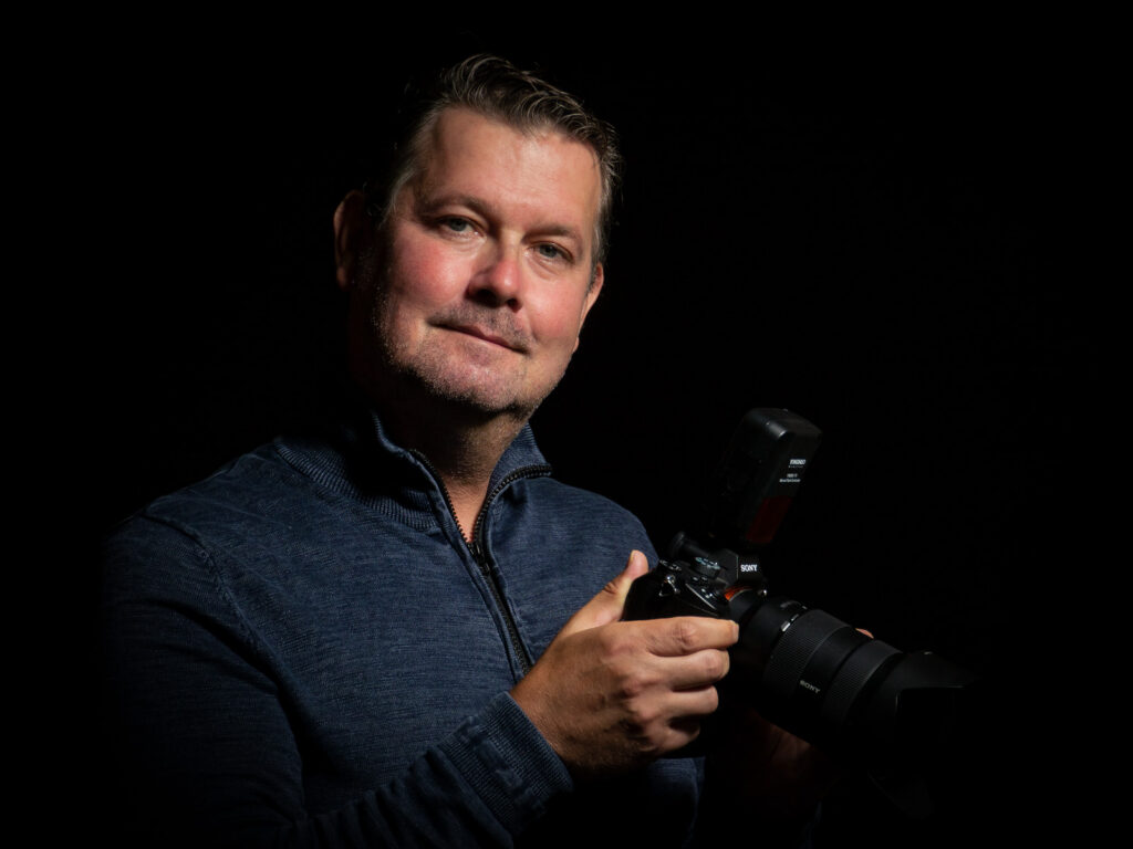 Fotograf Mario Hambsch mit Kamera in der Hand vor dunklem Hintergrund